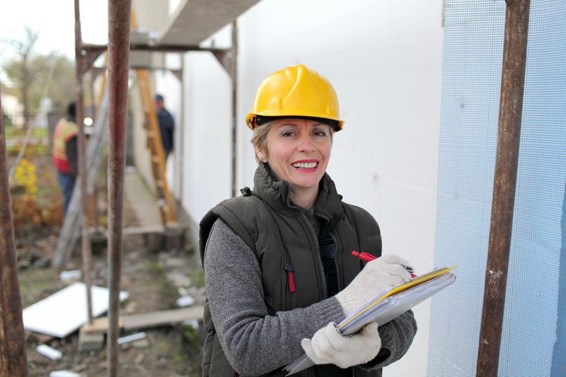 照片中，一位戴着黄色安全帽、穿着灰色毛衣和黑色外套背心、戴着白手套、面带微笑的妇女在黄色的写字板上写着什么，建筑工人在后面干活。